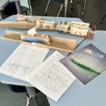 Model trains made from consumables on a desk with 2 completed pages of student design and reflection work.